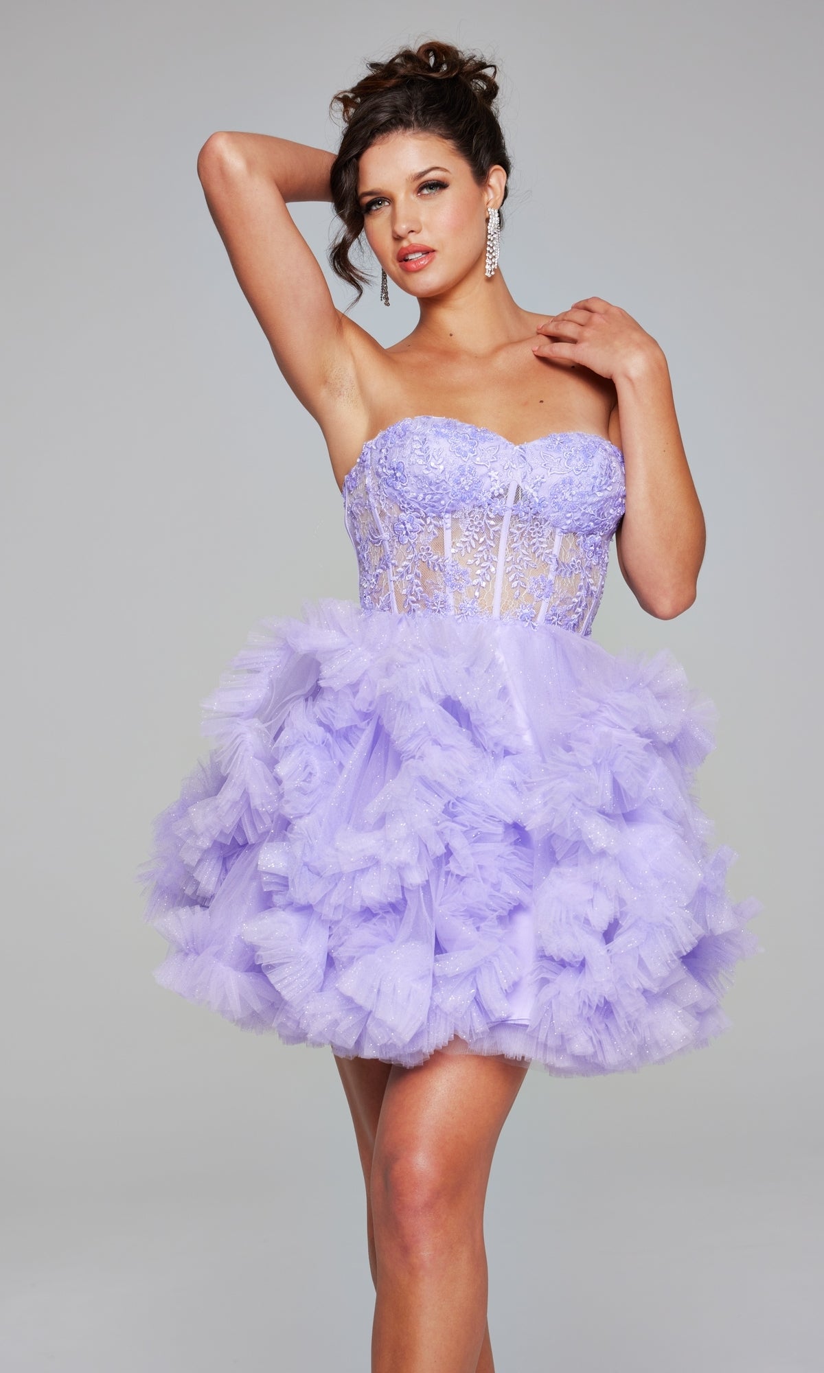 A woman poses with one arm raised, wearing the Short Homecoming Dress 40628 by Jovani, featuring a strapless lilac mini design with a sheer lace corset bodice and a voluminous ruffled tulle skirt.